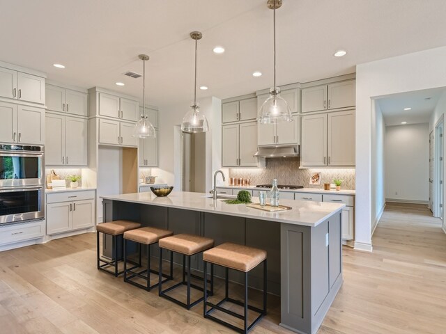 kitchen featuring hanging light fixtures, appliances with stainless steel finishes, an island with sink, tasteful backsplash, and light hardwood / wood-style flooring