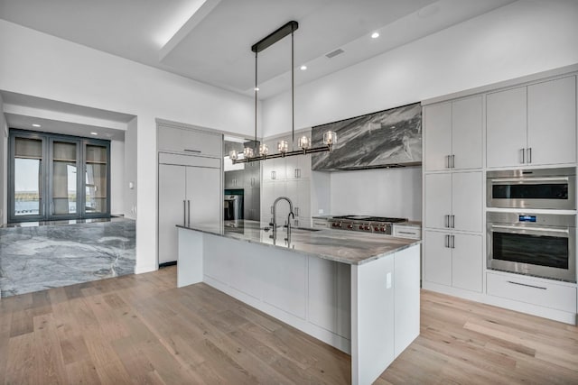 kitchen with wall chimney range hood, light hardwood / wood-style floors, sink, and a center island with sink