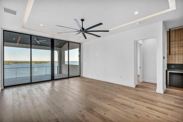 unfurnished living room with light wood-type flooring, a water view, ceiling fan, and a tray ceiling