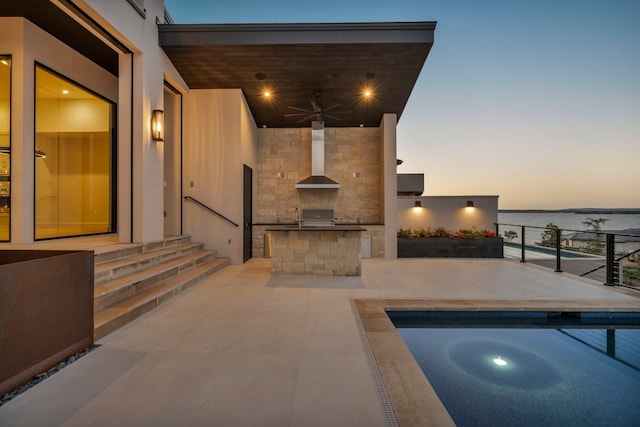 patio terrace at dusk with an outdoor kitchen, a pool, sink, a water view, and ceiling fan