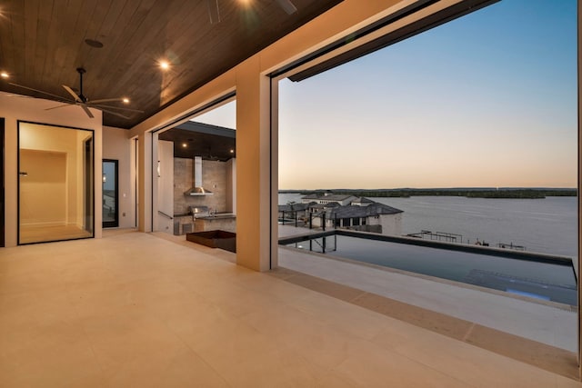 patio terrace at dusk with a water view, ceiling fan, a balcony, and exterior kitchen