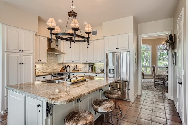 kitchen featuring wall chimney range hood, backsplash, stainless steel appliances, hanging light fixtures, and a center island with sink