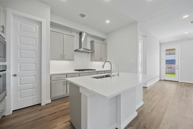 kitchen with gas cooktop, sink, wall chimney range hood, light hardwood / wood-style floors, and an island with sink