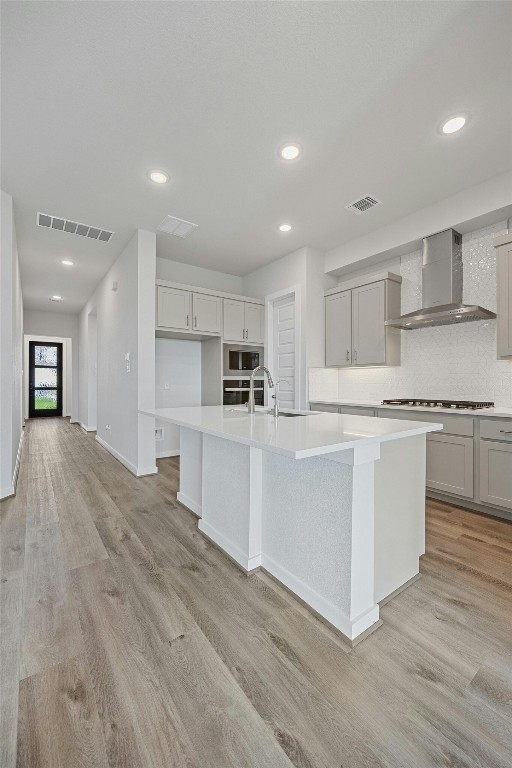 kitchen featuring sink, wall chimney exhaust hood, light wood-type flooring, a center island with sink, and appliances with stainless steel finishes