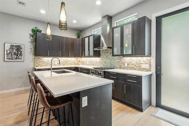 kitchen featuring hanging light fixtures, tasteful backsplash, a kitchen island with sink, wall chimney exhaust hood, and light wood-type flooring