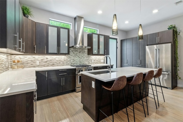 kitchen featuring wall chimney range hood, backsplash, high end appliances, a kitchen island with sink, and light wood-type flooring