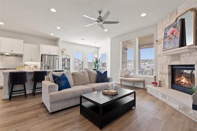 living room featuring ceiling fan, a stone fireplace, a textured ceiling, and light hardwood / wood-style floors