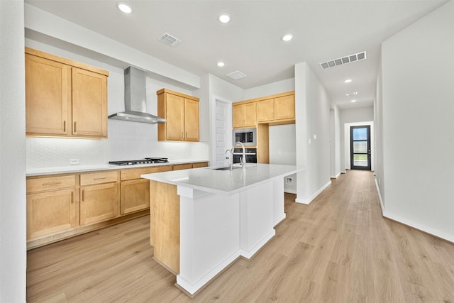 kitchen with wall chimney exhaust hood, backsplash, light wood-type flooring, a center island with sink, and appliances with stainless steel finishes