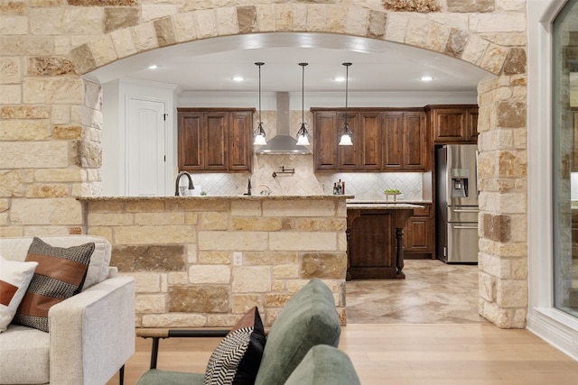 kitchen with light stone counters, ornamental molding, wall chimney range hood, pendant lighting, and stainless steel fridge with ice dispenser