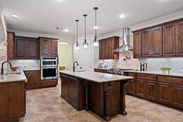 kitchen with sink, an island with sink, wall chimney range hood, and appliances with stainless steel finishes