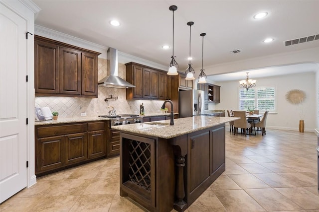 kitchen featuring wall chimney exhaust hood, stainless steel appliances, a kitchen island with sink, sink, and pendant lighting