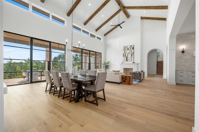 dining room featuring high vaulted ceiling, light wood-style flooring, ceiling fan with notable chandelier, a fireplace, and beamed ceiling