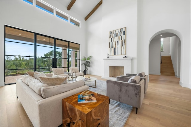 living room featuring arched walkways, a fireplace with raised hearth, light wood-style flooring, a high ceiling, and beam ceiling