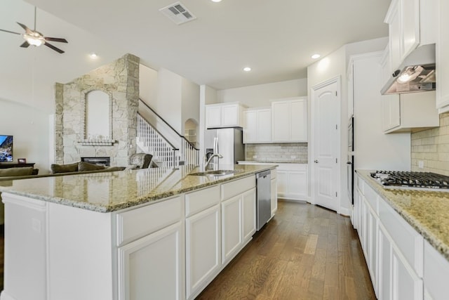 kitchen with a fireplace, a center island with sink, ceiling fan, dark wood-type flooring, and appliances with stainless steel finishes