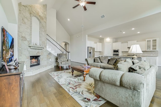 living room with high vaulted ceiling, a stone fireplace, ceiling fan, and light wood-type flooring