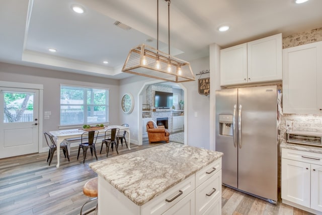 kitchen with a brick fireplace, a tray ceiling, pendant lighting, stainless steel fridge with ice dispenser, and white cabinetry