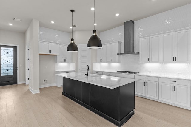 kitchen with wall chimney range hood, gas cooktop, white cabinets, and a center island with sink