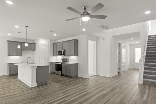 kitchen with ceiling fan, a kitchen island with sink, stainless steel appliances, gray cabinetry, and dark wood-type flooring