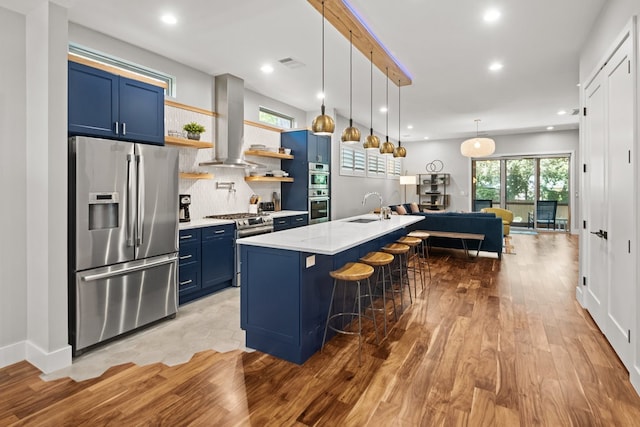 kitchen featuring a center island with sink, wall chimney exhaust hood, blue cabinetry, appliances with stainless steel finishes, and a breakfast bar