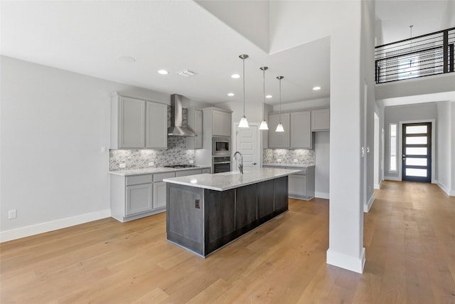 kitchen with appliances with stainless steel finishes, wall chimney range hood, an island with sink, and light hardwood / wood-style flooring