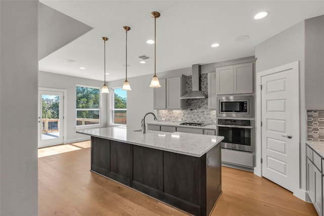 kitchen featuring a center island with sink, stainless steel appliances, light hardwood / wood-style flooring, wall chimney exhaust hood, and tasteful backsplash