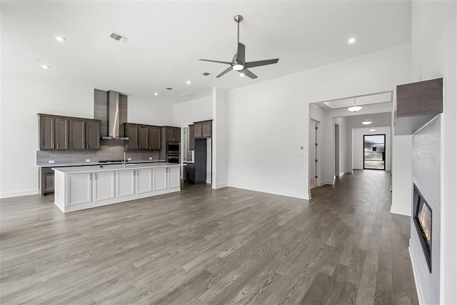 kitchen featuring appliances with stainless steel finishes, wall chimney exhaust hood, ceiling fan, a center island with sink, and light hardwood / wood-style flooring