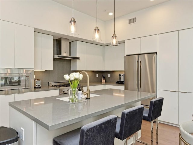 kitchen featuring a center island with sink, appliances with stainless steel finishes, hanging light fixtures, white cabinets, and wall chimney range hood