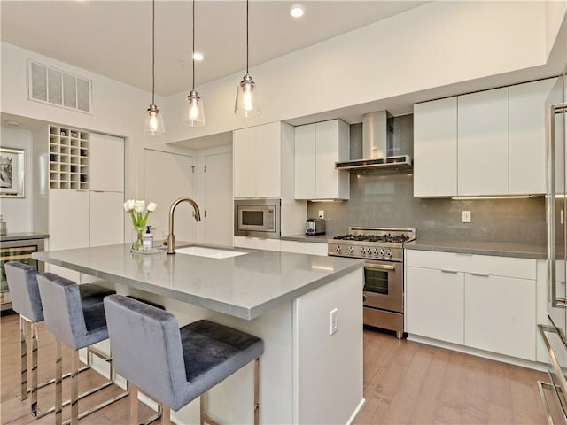 kitchen with sink, stainless steel appliances, wall chimney range hood, and a center island with sink