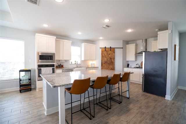 kitchen featuring wall chimney range hood, white cabinetry, appliances with stainless steel finishes, a center island, and a barn door