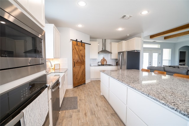 kitchen with white cabinets, wall chimney range hood, stainless steel appliances, light wood-type flooring, and a barn door