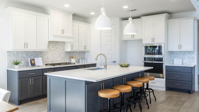 kitchen with stainless steel appliances, sink, white cabinetry, hanging light fixtures, and an island with sink