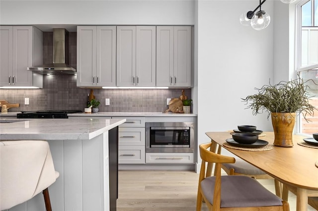 kitchen featuring stainless steel microwave, gray cabinetry, backsplash, light stone counters, and wall chimney exhaust hood