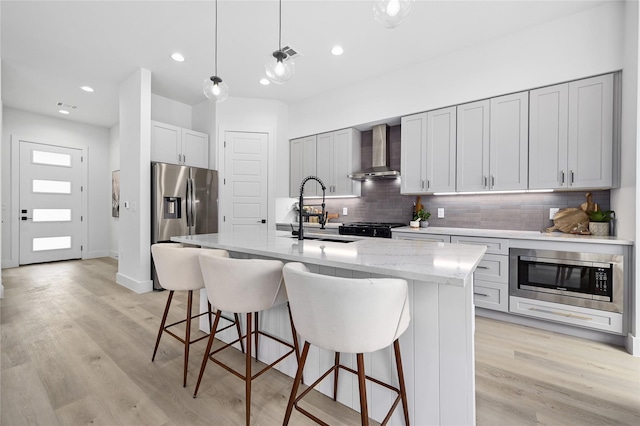 kitchen with sink, wall chimney range hood, hanging light fixtures, stainless steel appliances, and a center island with sink