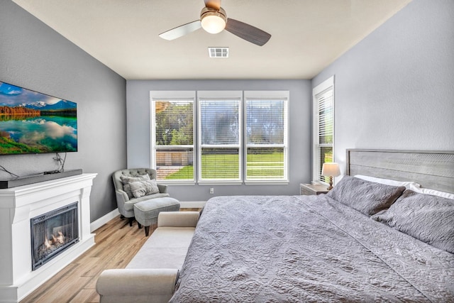 bedroom with visible vents, a ceiling fan, a glass covered fireplace, wood finished floors, and baseboards