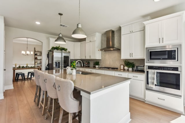 kitchen with wall chimney exhaust hood, light hardwood / wood-style floors, a center island with sink, sink, and stainless steel appliances