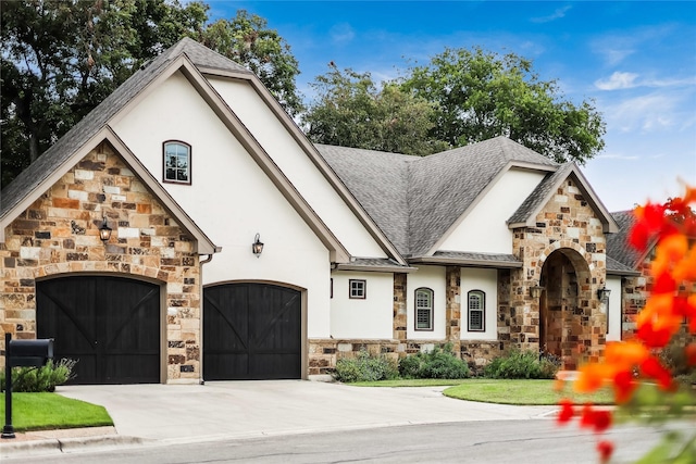 french country inspired facade with a playground, stucco siding, a shingled roof, an attached garage, and driveway