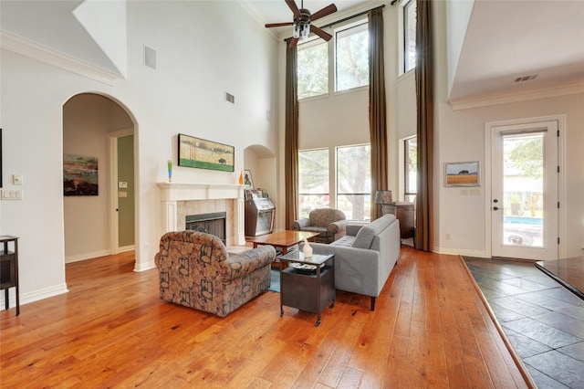 living room featuring a towering ceiling, light wood-type flooring, ceiling fan, crown molding, and a fireplace