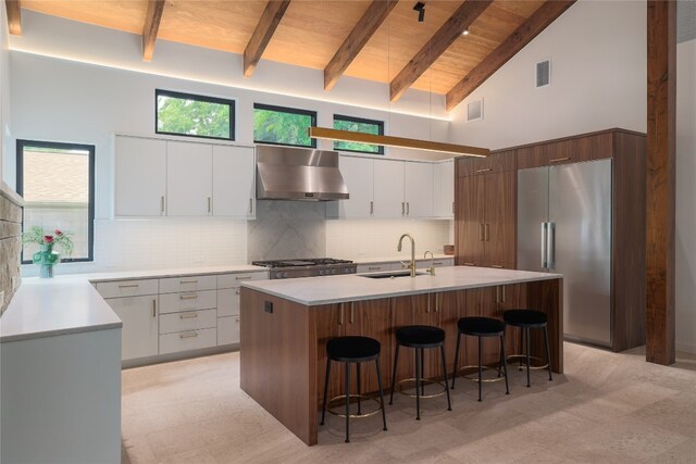 kitchen featuring wall chimney range hood, a kitchen island with sink, beam ceiling, sink, and tasteful backsplash