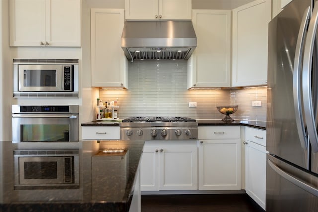 kitchen with dark stone countertops, range hood, backsplash, white cabinets, and appliances with stainless steel finishes