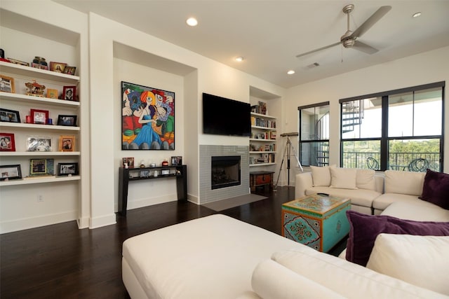 living room with built in features, a tiled fireplace, ceiling fan, and dark wood-type flooring