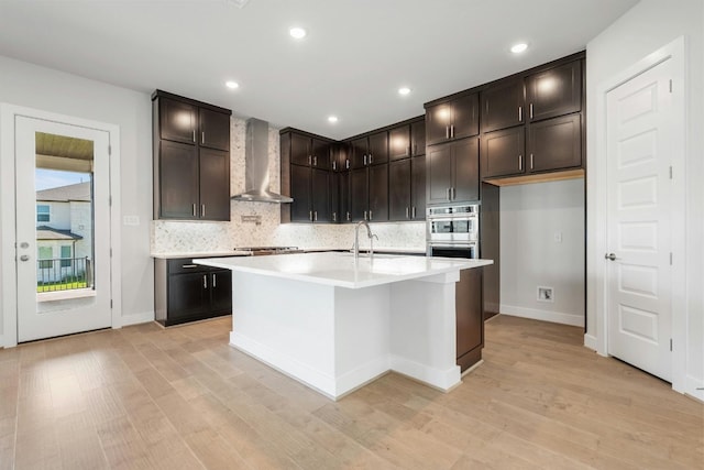 kitchen featuring decorative backsplash, wall chimney exhaust hood, light countertops, light wood-type flooring, and a sink