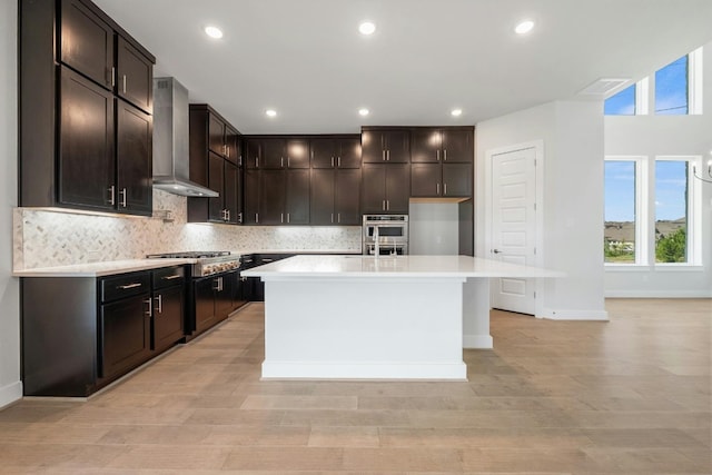 kitchen with wall chimney range hood, dark brown cabinets, a center island with sink, and stainless steel appliances