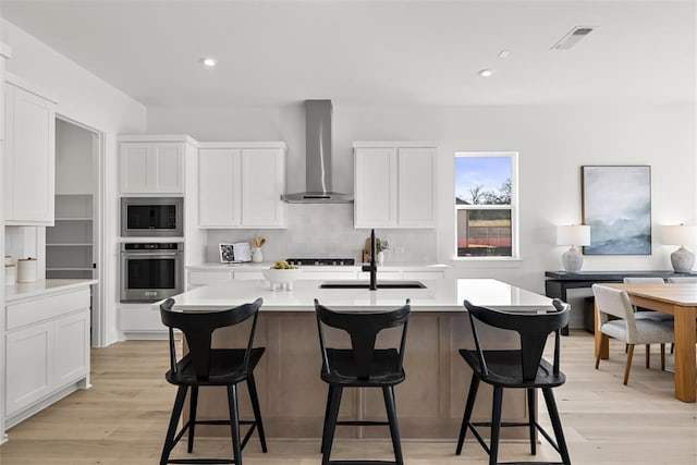 kitchen featuring appliances with stainless steel finishes, light wood-type flooring, wall chimney range hood, a center island with sink, and white cabinetry