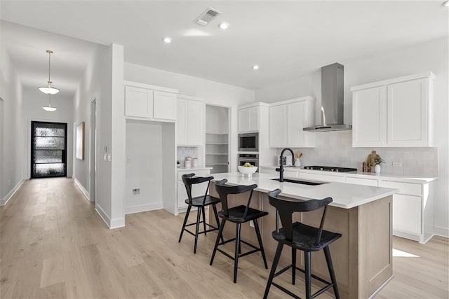 kitchen featuring wall chimney exhaust hood, sink, light hardwood / wood-style flooring, white cabinetry, and an island with sink