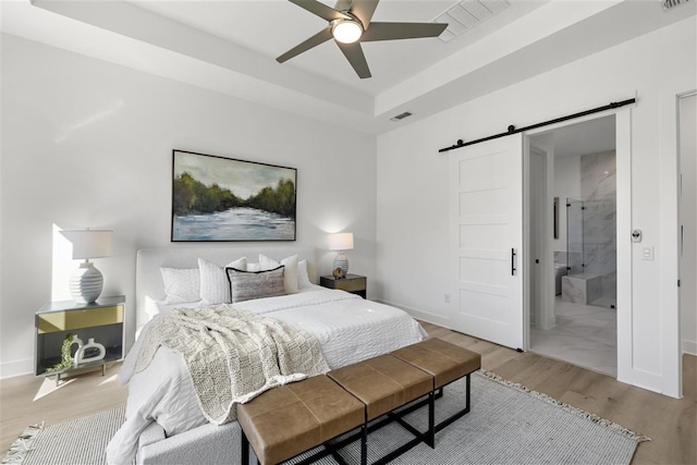 bedroom featuring ensuite bathroom, ceiling fan, a barn door, light wood-type flooring, and a tray ceiling