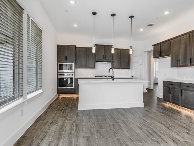 kitchen with appliances with stainless steel finishes, hanging light fixtures, dark brown cabinetry, dark wood-type flooring, and a kitchen island with sink