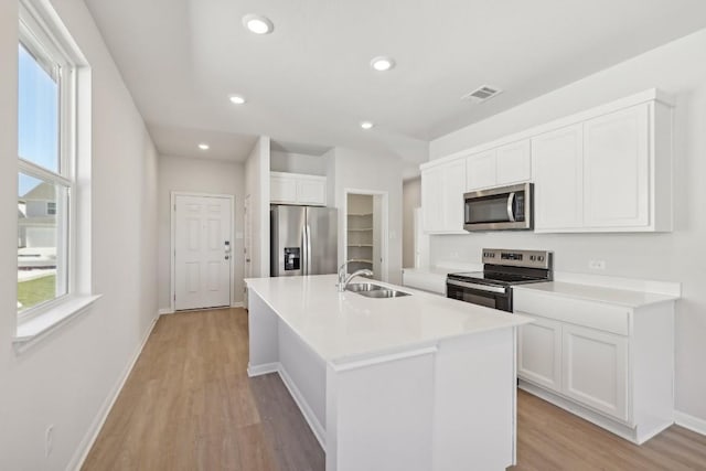 kitchen with sink, a kitchen island with sink, stainless steel appliances, white cabinets, and light wood-type flooring