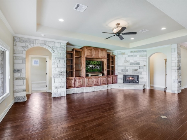 unfurnished living room with dark hardwood / wood-style flooring, ceiling fan, a raised ceiling, a stone fireplace, and ornamental molding