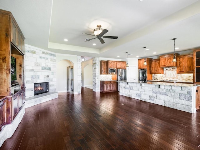 unfurnished living room with a tray ceiling, arched walkways, ceiling fan, dark wood-style floors, and a tiled fireplace