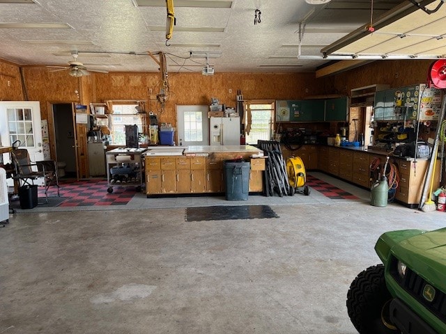 garage featuring a workshop area, ceiling fan, wood walls, and white refrigerator with ice dispenser